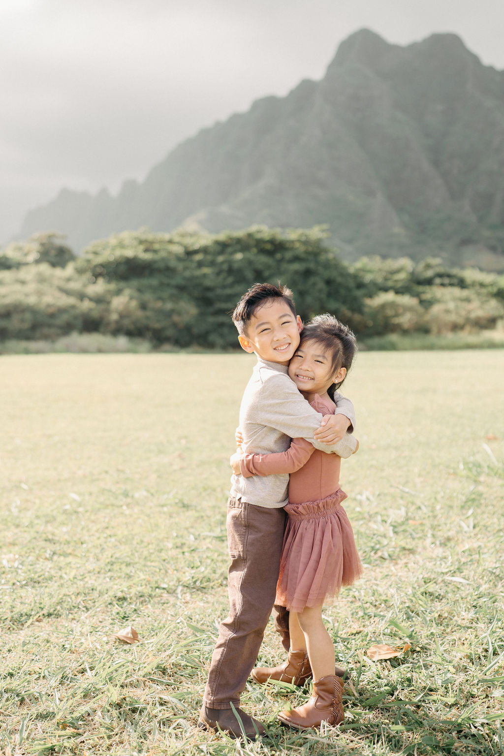 Two young children, a boy and a girl, standing on grass hug each other and smile at the camera with green trees and mountains in the background.