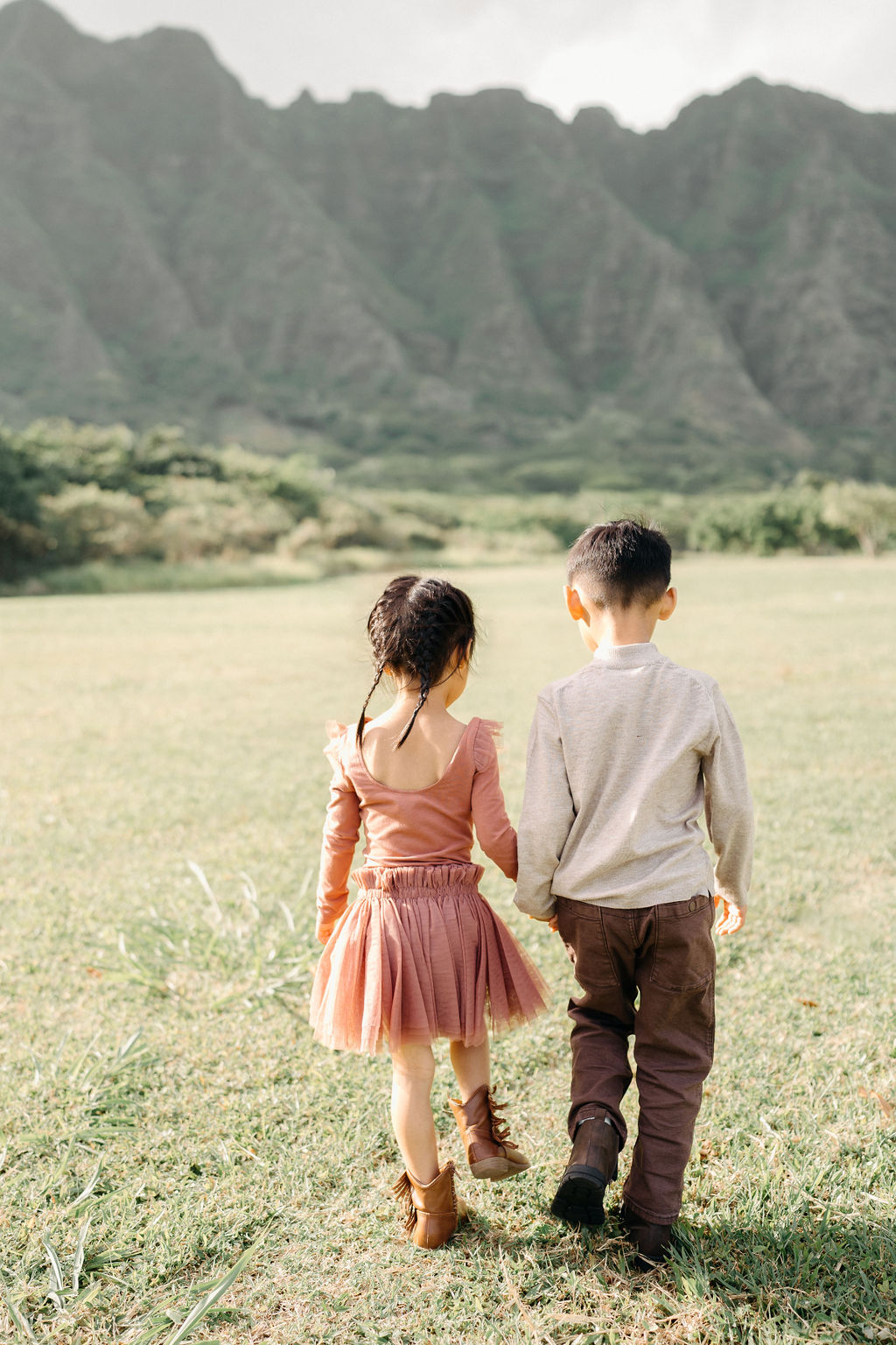Two young children, a boy and a girl, standing on grass hug each other and smile at the camera with green trees and mountains in the background.