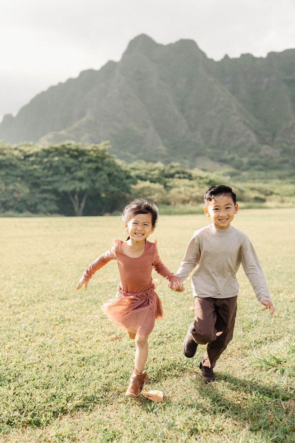 Two young children, a boy and a girl, standing on grass hug each other and smile at the camera with green trees and mountains in the background.