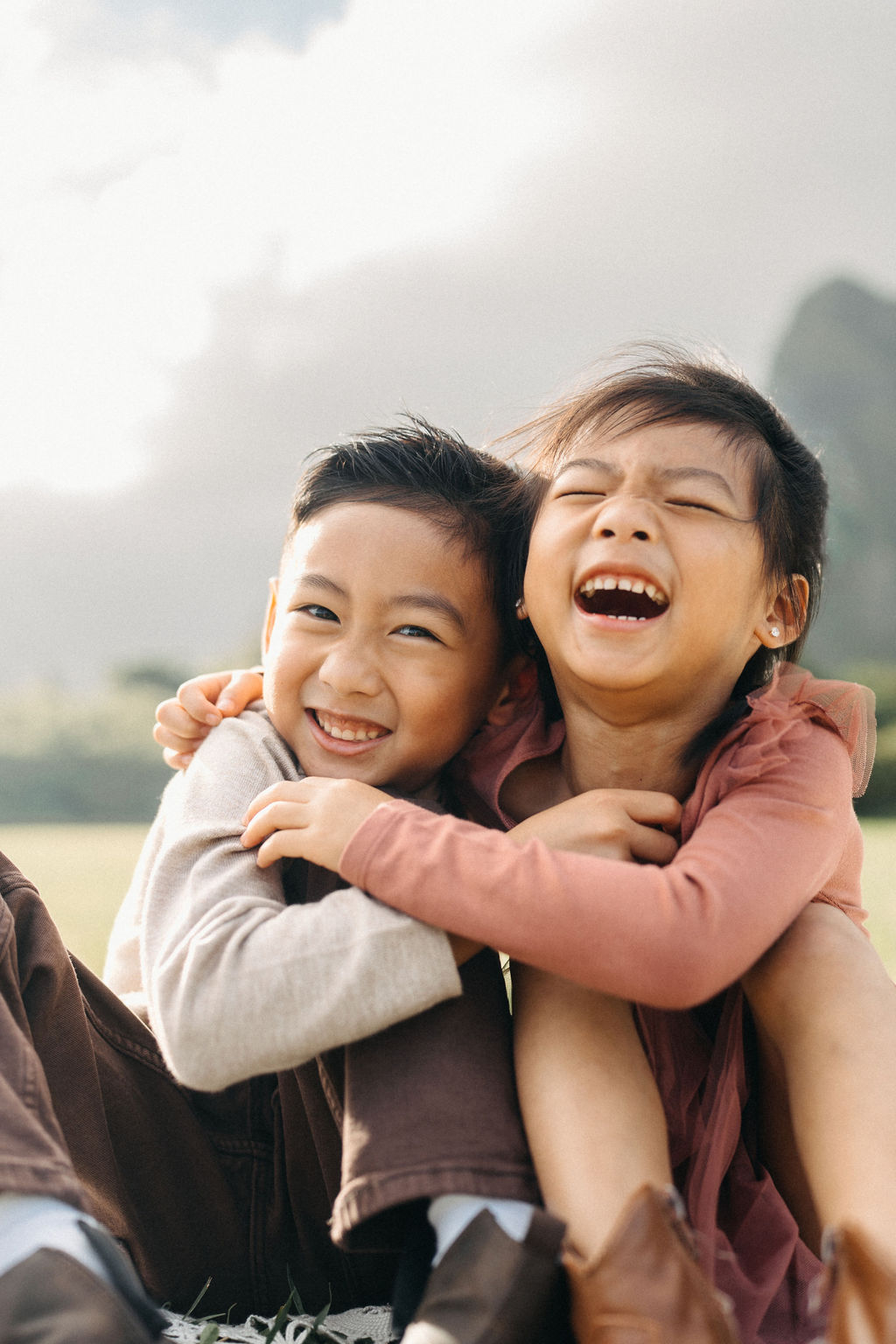 Two young children, a boy and a girl, stand outside on grass and smile while hugging each other. Trees and greenery are blurred in the background.