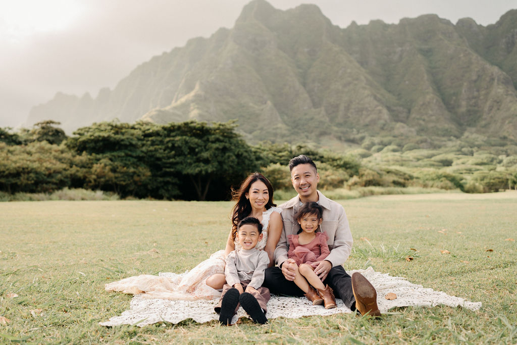 A family of four poses together on a grassy field with green mountains in the background for their mini family session