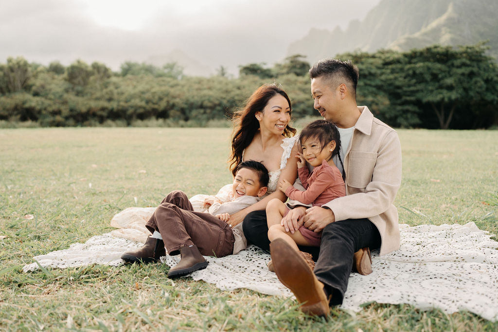 A family of four poses together on a grassy field with green mountains in the background for their mini family session