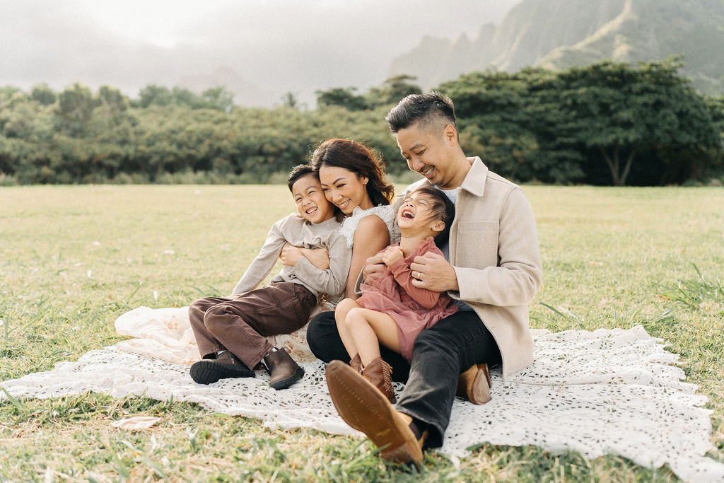 A family of four poses together on a grassy field with green mountains in the background