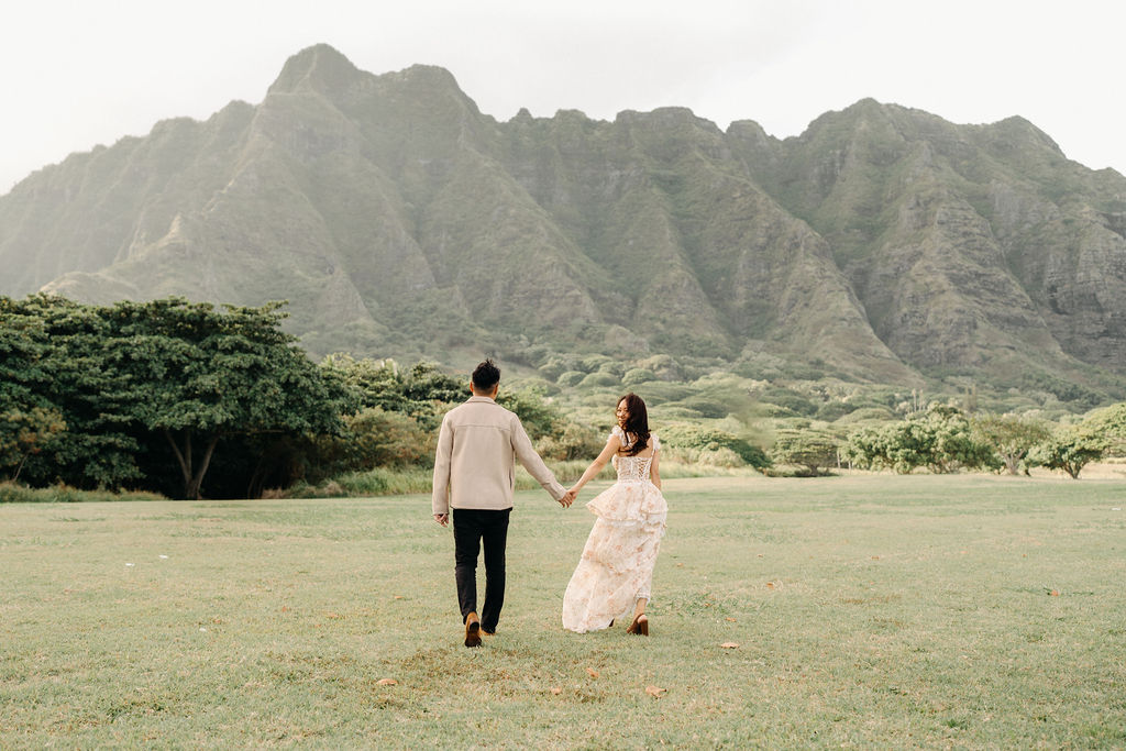 A couple stands embracing in a grassy field with mountains and trees in the background under a cloudy sky.
