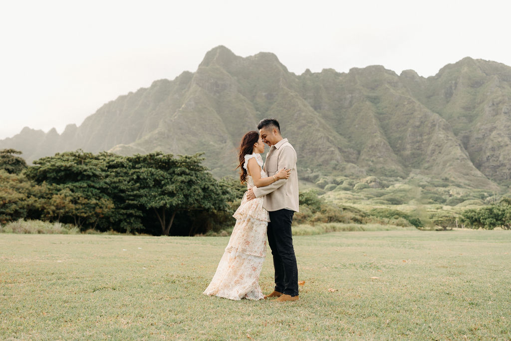 A couple stands embracing in a grassy field with mountains and trees in the background under a cloudy sky.
