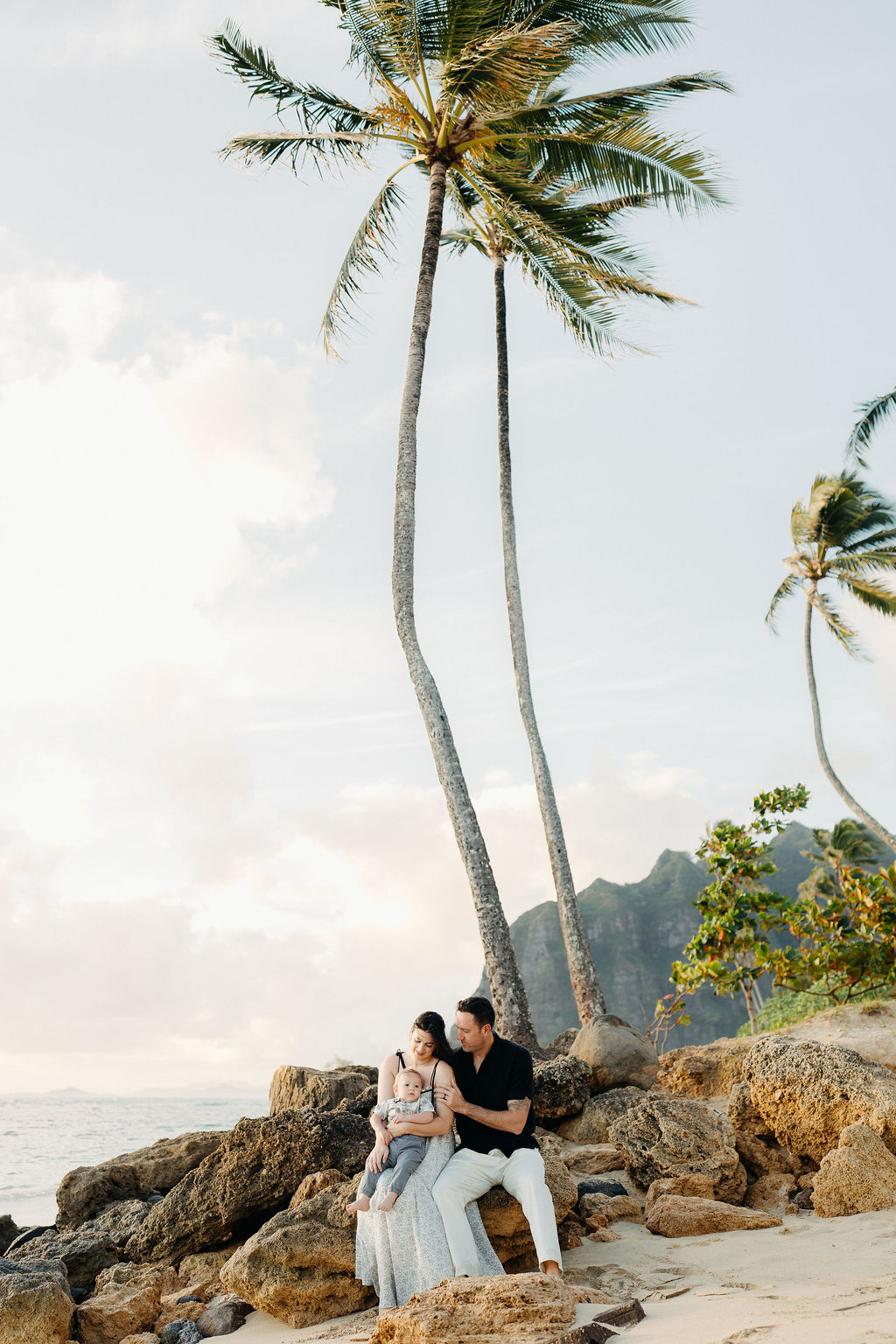 A man, woman, and young child walk barefoot along a sandy beach with mountains and palm trees in the background.