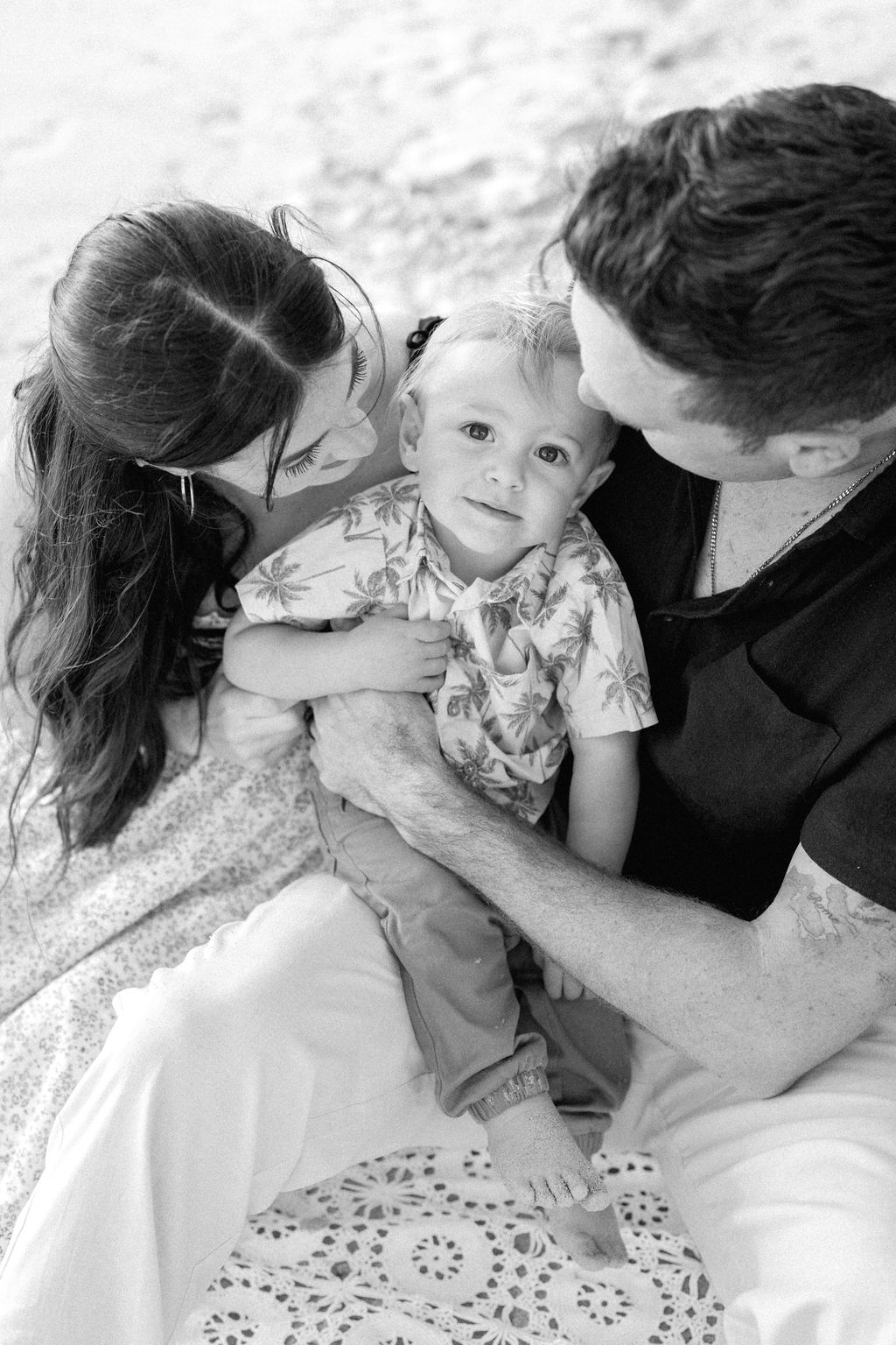 A man and woman sit closely together on a blanket at the beach, holding a young child between them. The image is in black and white.