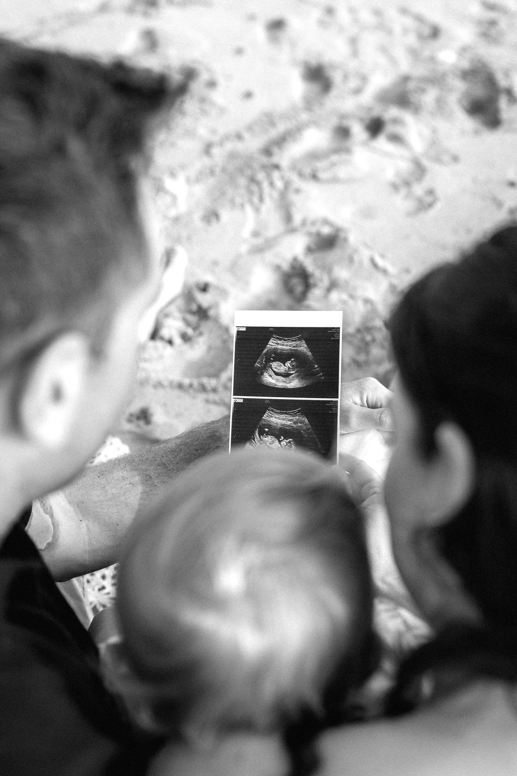 A man, woman, and child look at an ultrasound photo, viewed from behind, with a sandy ground visible in the background.