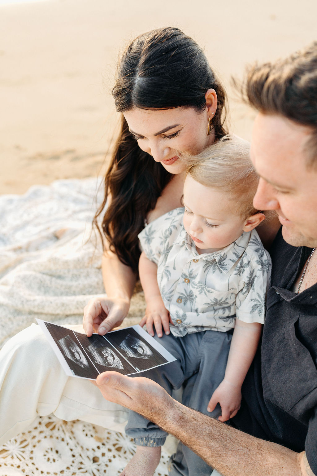 A family of three sits on a sandy beach near the ocean, with palm trees and mountains in the background. The parents smile as they lift their child into the air.