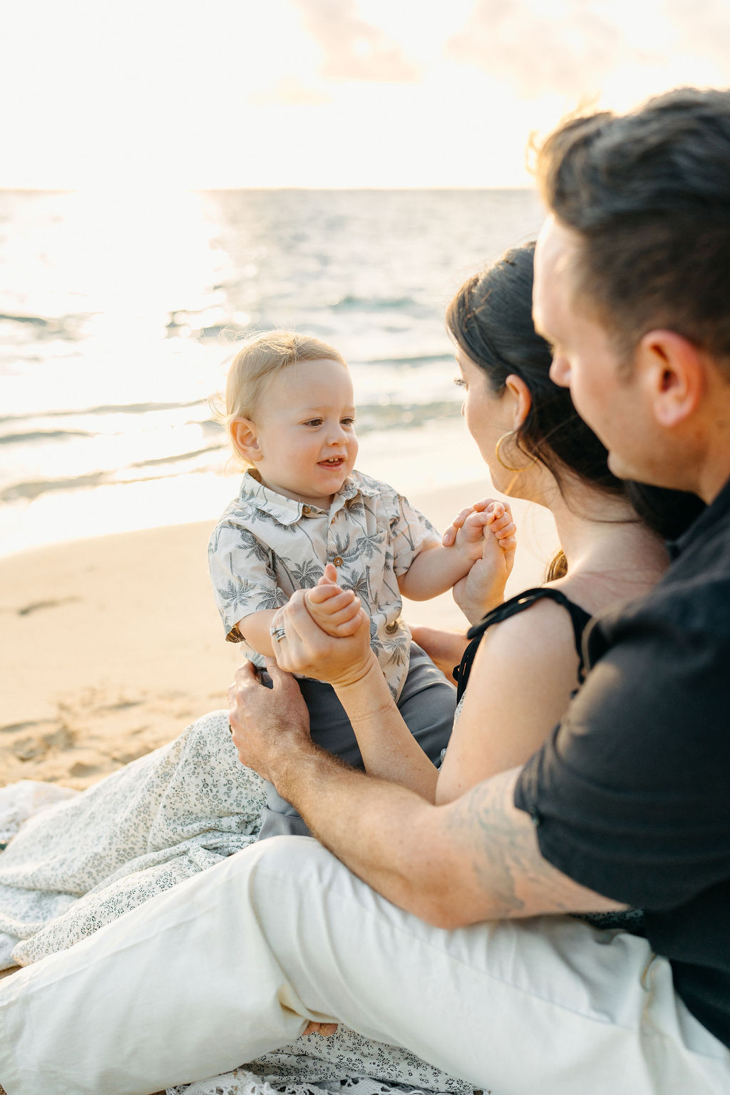 A family of three sits on a sandy beach near the ocean, with palm trees and mountains in the background. The parents smile as they lift their child into the air.