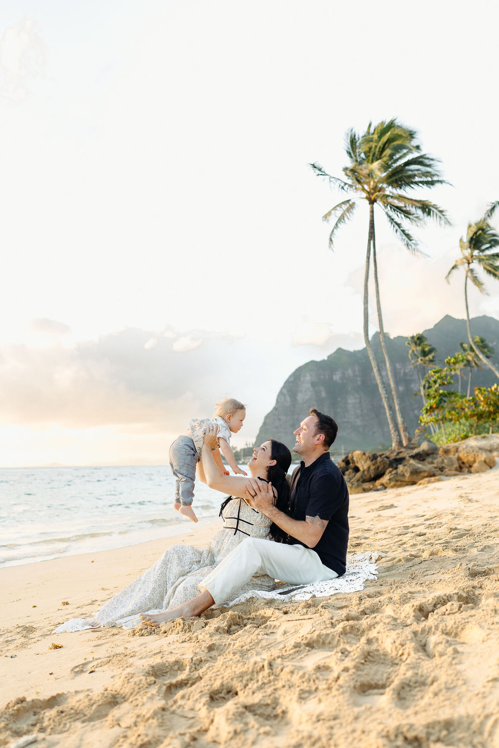 A family of three sits on a sandy beach near the ocean, with palm trees and mountains in the background. The parents smile as they lift their child into the air.