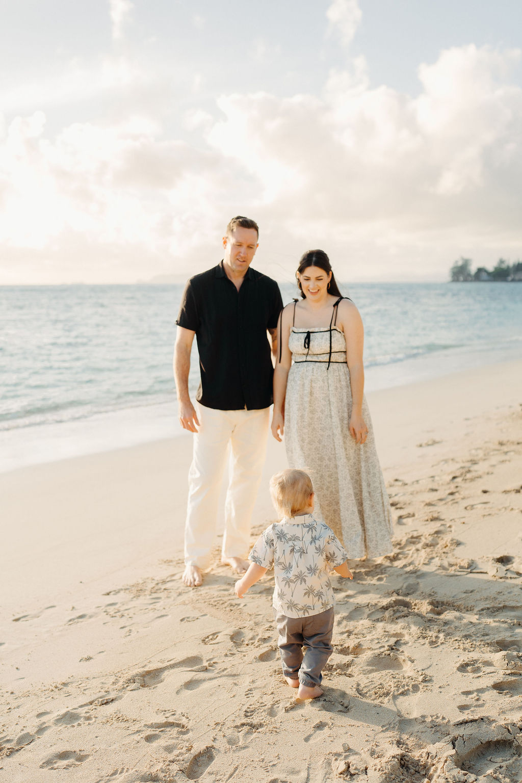 A man and woman stand together on a sandy beach while a small child walks toward them, with the ocean and sky in the background.