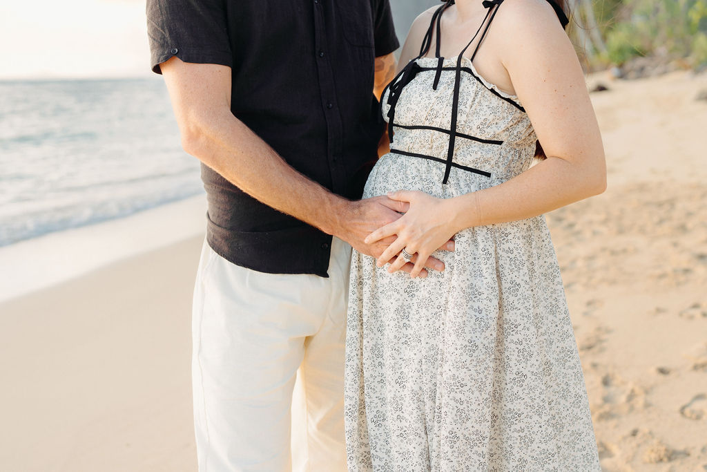 A couple stands on a sandy beach, with the man gently holding the woman's pregnant belly.